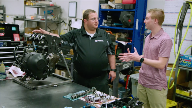 Two men looking at and talking about the engine that goes in a Legend car