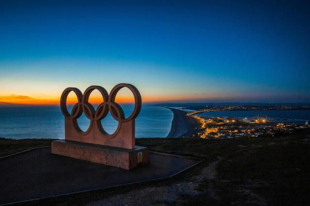 Olympic rings monument on a hill overlooking a coastal city during sunset with the coastline stretching into the distance.