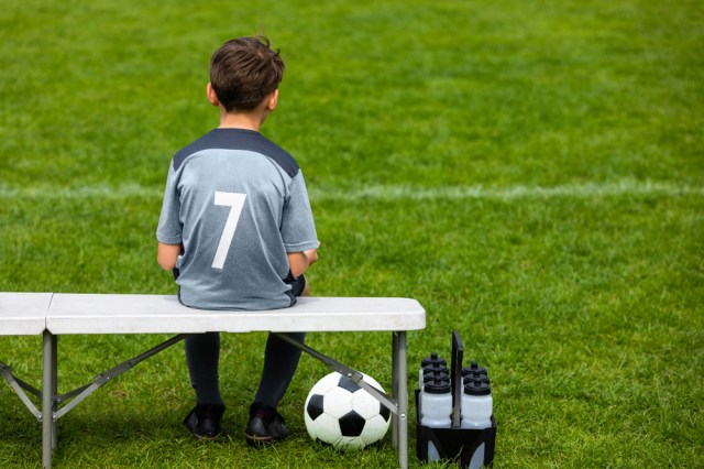 Little soccer player sitting on a wooden bench and watching soccer game. Young substitute player waiting on a soccer bench.