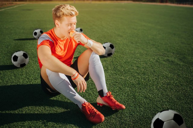 Hungry young player sitting on lawn and eating apple