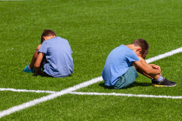 education, bullying, conflict, social relations and people concept - two sad disappointed boys sitting back to back on the grass in stadium