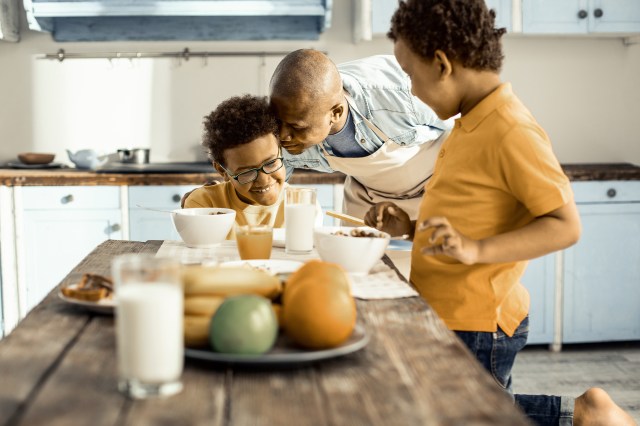Delicate man in apron in turns sincerely hugging his sons in the kitchen while they cook.