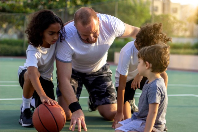 Shot of three young boys and their coach chatting outside on basketball court.