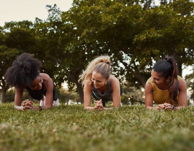 Three multiethnic smiling young active and sporty women doing plank exercises in the park - three woman being active outdoors