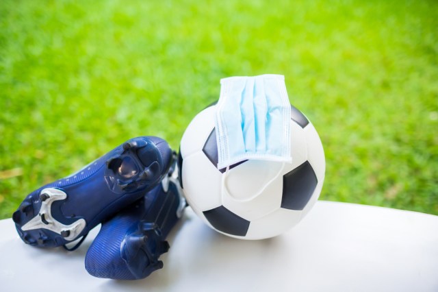 Mask is on football with soccer sport shoes on white floor with green blur background. The concept of Covid-19 impact to football matches.