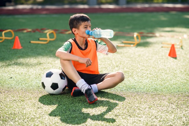 Tired teen boy in soccer uniform drinks with water from plastic bottle after intensive training at stadium in morning.