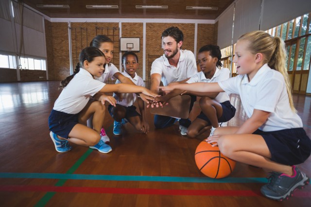 Sports teacher and school kids forming hand stack in basketball court at school gym