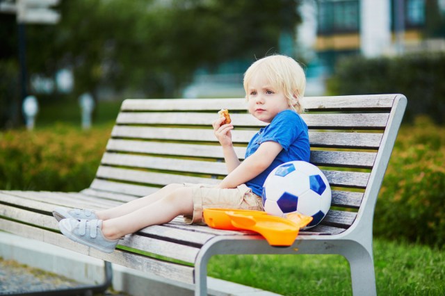Adorable little boy sitting on the bench with lunchbox in park