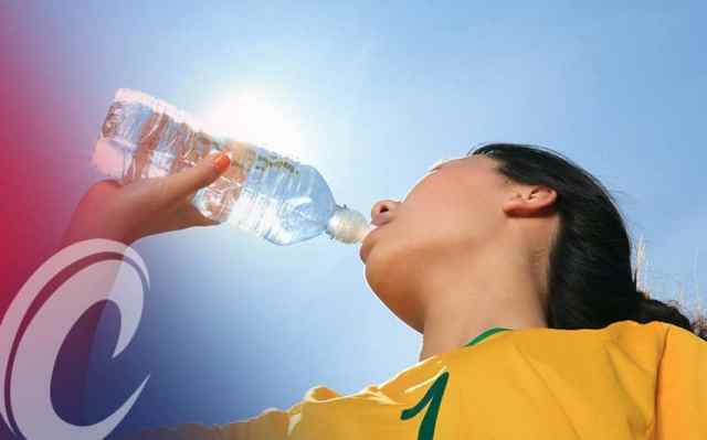Young female soccer player drinking a clear bottle of water against blue sky and bright sun.